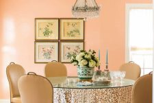 refined Peach Fuzz dining room with a table with a glass tabletop, beige chairs, a crystal chandelier and a gallery wall