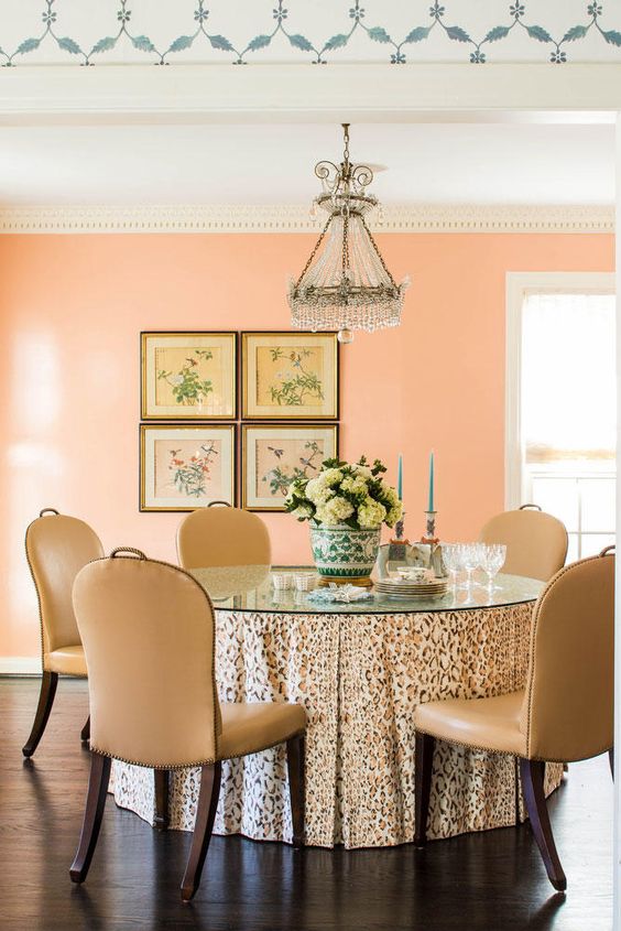 refined Peach Fuzz dining room with a table with a glass tabletop, beige chairs, a crystal chandelier and a gallery wall