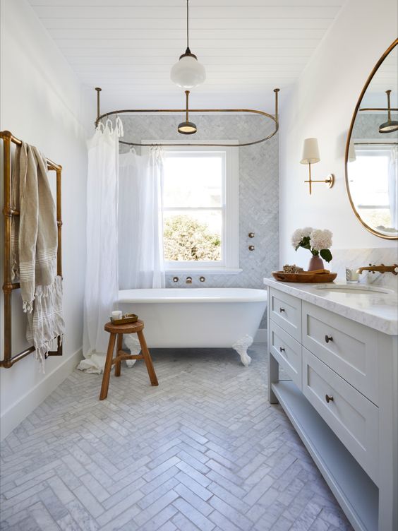 a beautiful and airy bathroom clad with white marble herringbone tiles, a large vanity, a vintage tub and brass fixtures