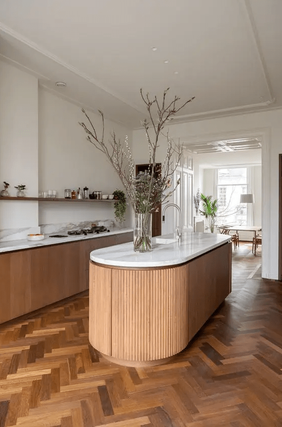 A beautiful and chic kitchen with a refined herringbone floor, light stained cabinets, a curved kitchen island and an open shelf