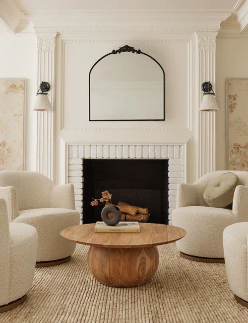 a beautiful and chic living room with a fireplace, creamy boucle chairs, a coffee table, a mirror and wall lamps