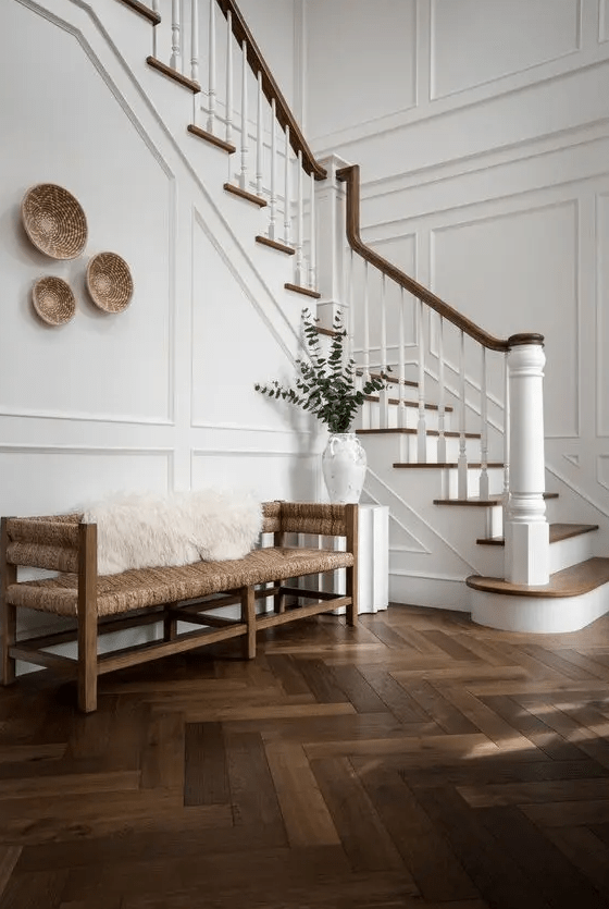 a beautiful and welcoming creamy entryway with white molded walls and a dark herringbone floor, a woven bench and some baskets