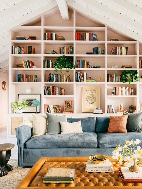 a beautiful living room with built-in bookshelves with blush backing, a blue sofa with pillows, an orange ottoman and greenery