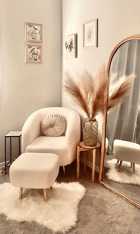 a booh nook with a mirror in a gold frame,a  boucle chair and a footrest, some art, layered rugs and pampas grass in the vase