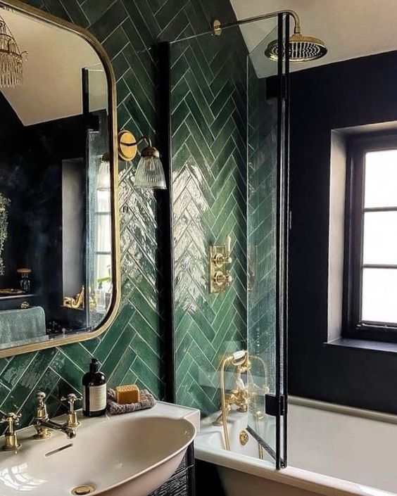 a chic bathroom done with green herringbone tiles, a tub with a glass screen and a vintage sink plus gold fixtures