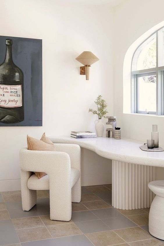 a contemporary home office with a neutral curved desk, a creamy boucle chair, a large artwork and a wall lamp