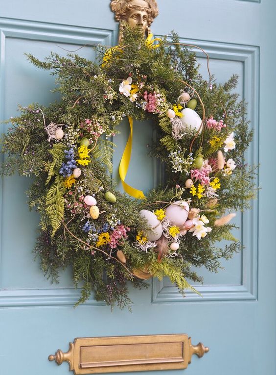 a gorgeous lush spring wreath with lots of foliage and greenery, yellow, blue and pink spring blooms, twigs, eggs and ribbon