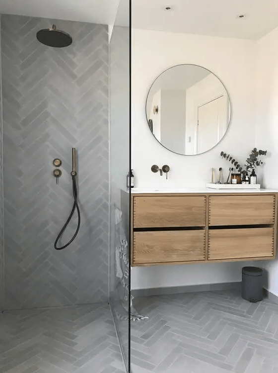 a modern bathroom done with grey herringbone tiles, a stained vanity, a round mirror and dark fixtures