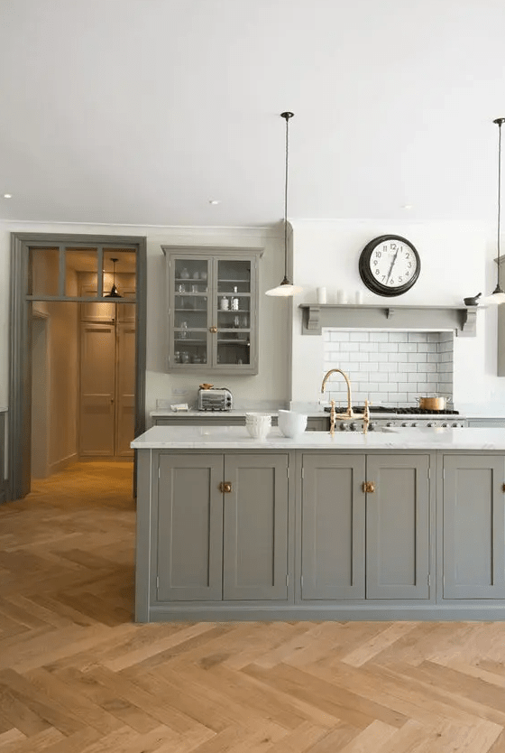 A modern farmhouse kitchen with white walls and a light stained herringbone floor, a grey kitchen island, a white backsplash and pendant lamps