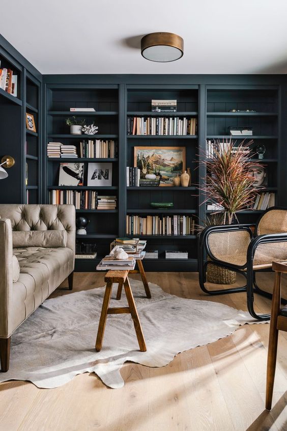 a navy home library with built-in shelves, a grey sofa, a rattan chair, a wooden bench and a rug and a grass