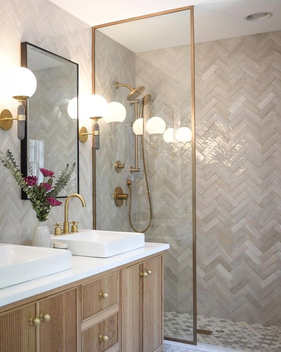 a neutral bathroom clad with tan and beige herringbone tiles, a shower space, a stained vanity and brass fixtures