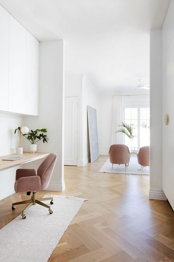 A neutral space with a herringbone floor and white walls, a built in desk and cabinets, pink chairs and lots of light