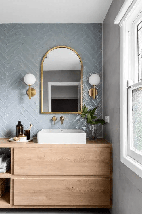 a pale blue herringbone tile wall, a stained vanity, a mirror with lamps and a white sink for a modern bathroom