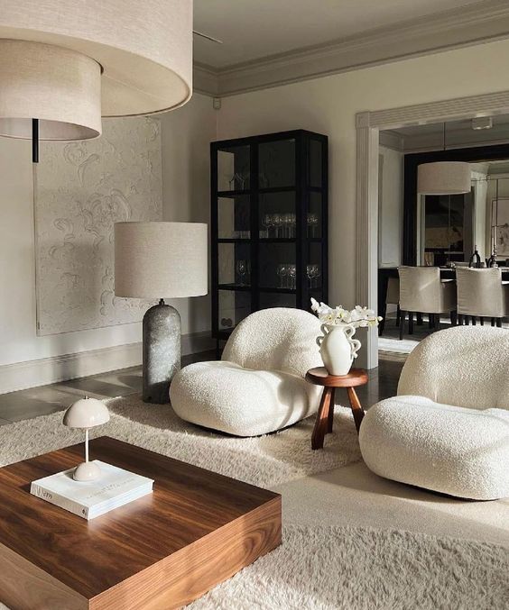 a refined living room with creamy curved boucle chairs, a coffee table,a  neutral rug, a storage unit and large floor lamps