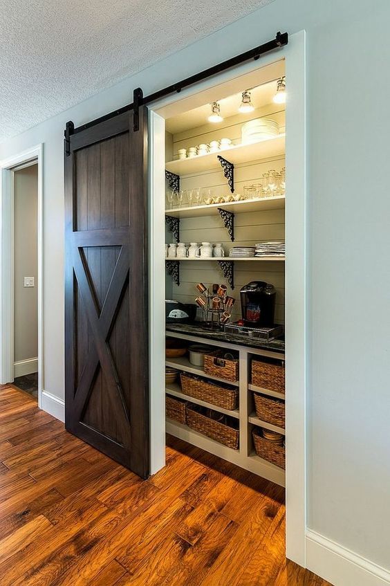 a rustic pantry with a dark stained barn door, open shelves and lights, baskets and various stuff for a rustic kitchen