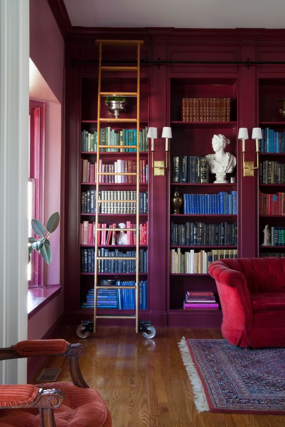 A sophisticated space with purple walls and built in shelves, a ladder, a red sofa, a printed rug and some chic decor