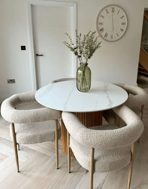 a stylish dining space with a round table with a marble tabletop and a fluted base, white boucle chairs, a clock and a green vase with greenery