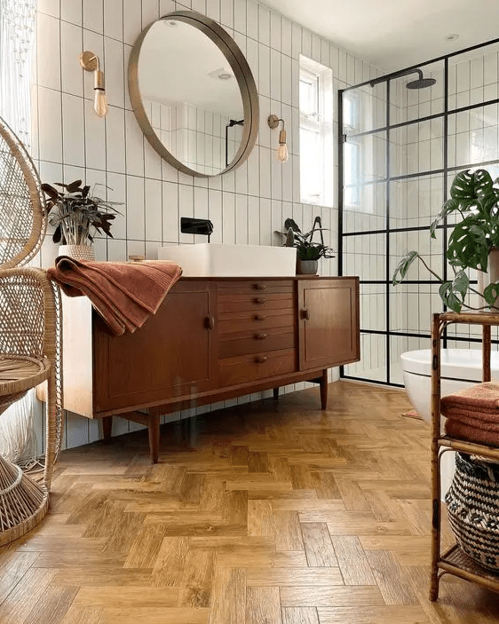 a stylish mid-century modern bathroom with white skinny tiles and a herringbone floor, a stained vanity, a papasan chair and a shower