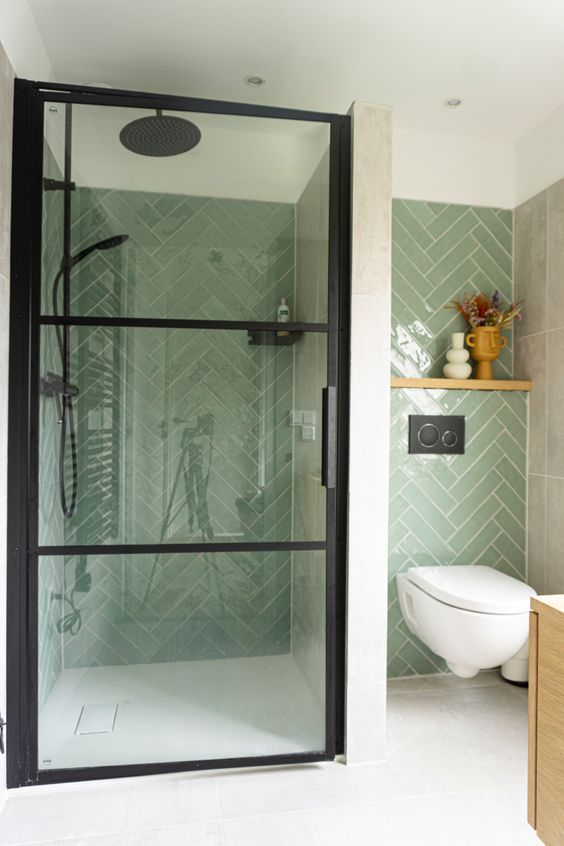 a stylish modern bathroom clad with light green herringbone tiles, a shower space and a floating toilet plus a stained vanity