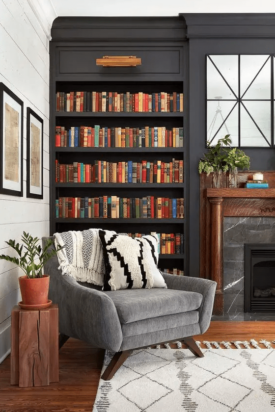 A stylish moody space with dark built in bookshelves, a marble fireplace and a grey chair for much comfort