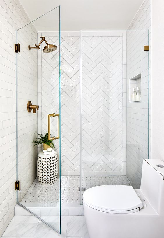 a stylish white bathroom with a shower space clad with herringbone tiles, a side table, some greenery and brass fixtures