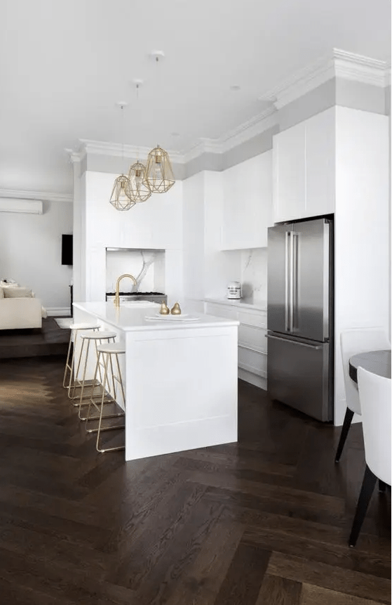 A white sleek kitchen with dark stained herringbone floors, gold touches and some built in appliances