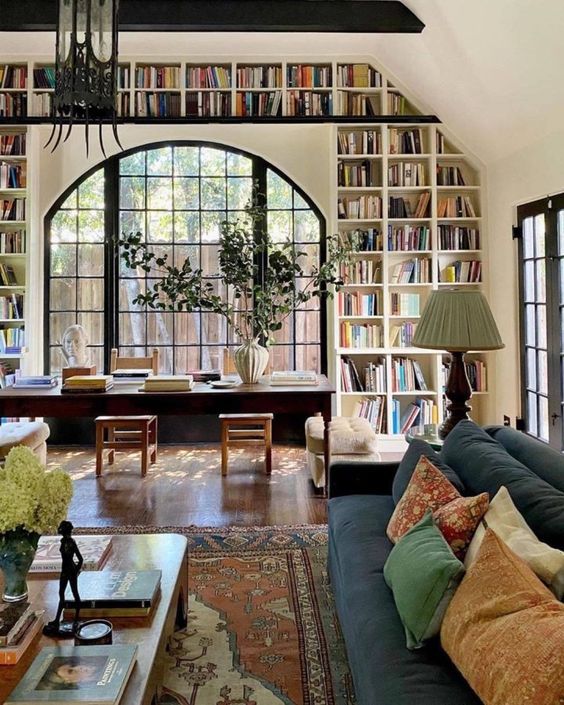 an airy living room with a large arched window, a bookshelves built in around it, eclectic furniture and lamps