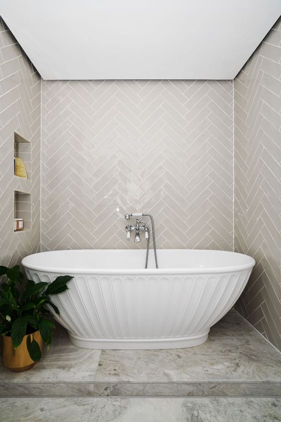 An eye catchy bathroom clad with grey herringbone tiles, with a textured tub and some niche shelves
