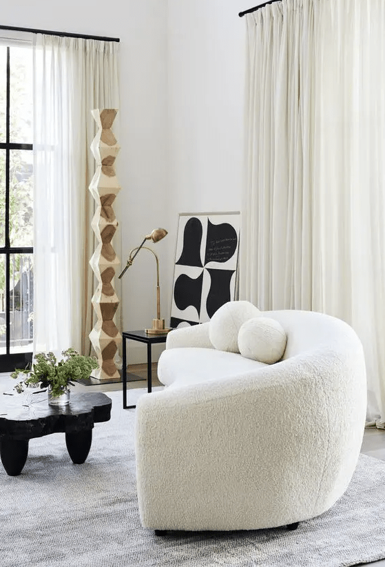 An eye catchy space with a white boucle curved sofa, a black tree slice table, a side table with a lamp and some abstract art
