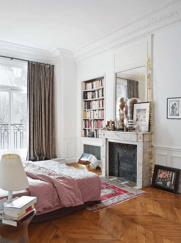 A French chic bedroom with paneling, an antique non working fireplace, a bed with pink bedding, built in shelves and a mirror