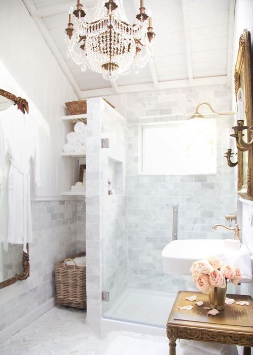 a French country bathroom with white marble tiles, a shower space, built-in shelves and a basket, a sink, a wooden table and a crystal chandelier
