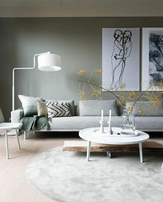 a Scandinavian living room with a green accent wall, a grey long sofa with pillows, some artwork and some white furniture and a lamp