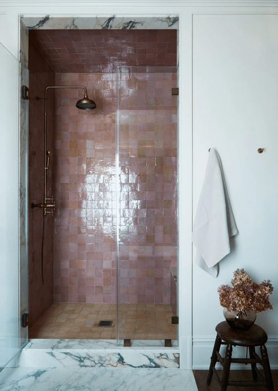a bathroom with a shower space clad with pink and tan Zellige tiles, brass fixtures and some blooms in a vase