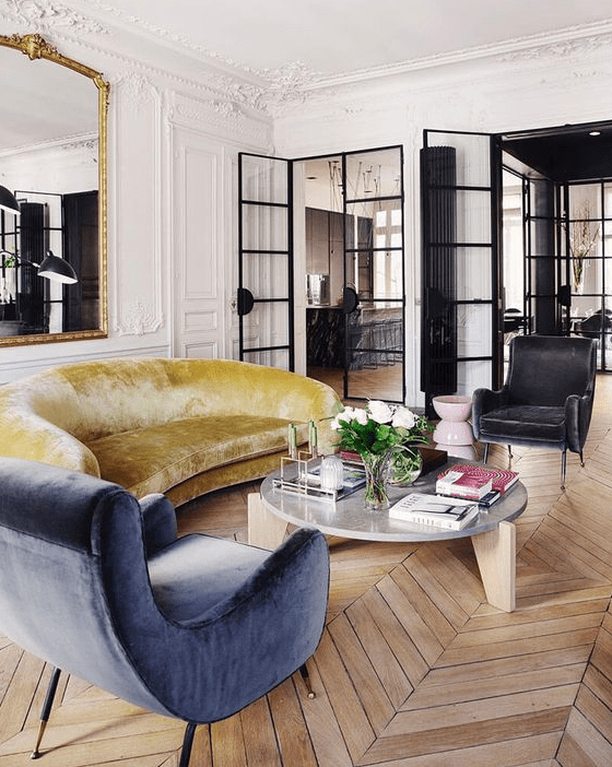 a beautiful Parisian living room with molding and chevron floors, a mustard sofa, a blue chair, a statement mirror in an ornated frame