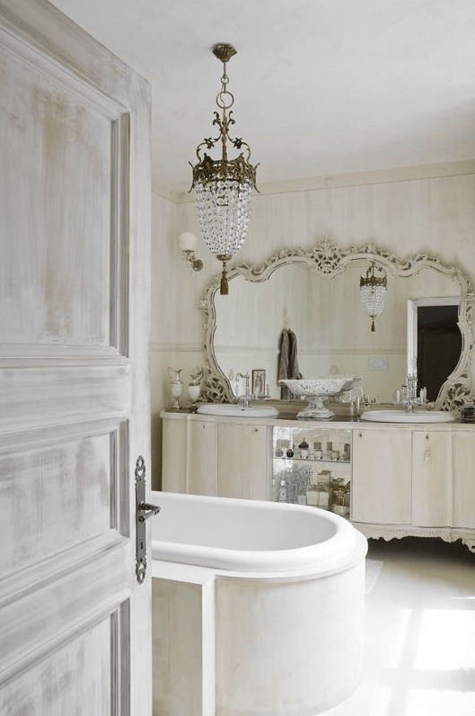 A beautiful Provence inspired bathroom with a large carved vanity and a mirror in a chic frame, a crystal pendant lamp and a modern tub