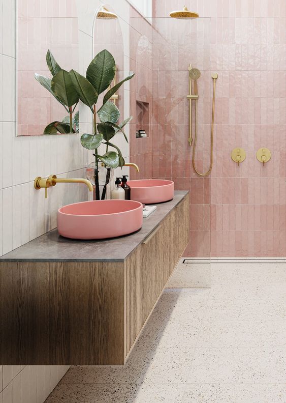 a beautiful modern bathroom with white stacked tiles and pink Zellige ones in the shower, a grey terrazzo floor, a stained vanity with pink sinks and gold hardware