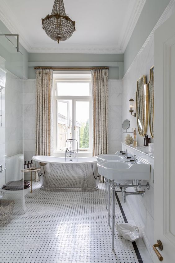 a beautiful neutral bathroom with white marble and printed tiles, a tub, a large crystal chandelier, sinks and mirrors