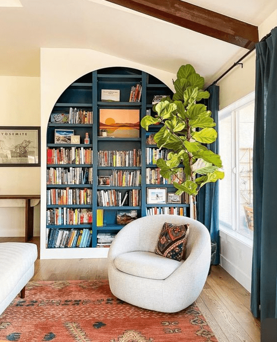 a beautiful reading nook with an arched bookcase with anvy backing and shelves, a creamy rounded chair and a bold rug