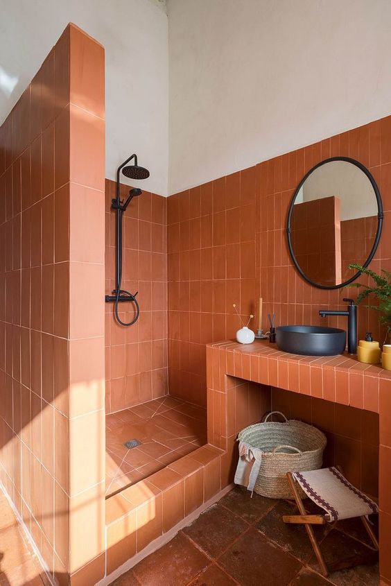 a bold terracotta tile bathroom with a vanity clad with tiles, a shower space, black hardware and a mirror in a frame