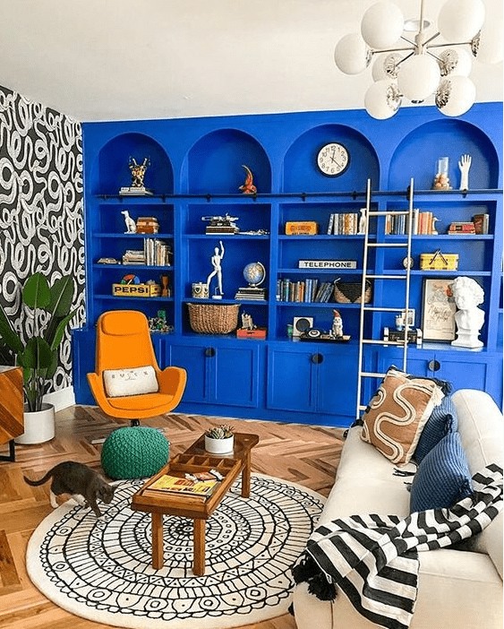 a bright maximalist space with bold blue arched bookcases, a white sofa and pillows, a printed rug, an orange chair and some decor