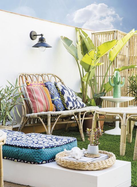 a bright patio with a green lawn, rattan furniture, a low bench with a cushion, potted plants, a wall lamp and a side table with a cactus lamp