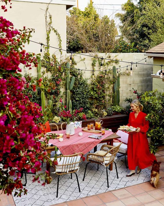 a bright patio with a planter with greenery and blooms, a table with rattan chairs, lights over the table and blooms around