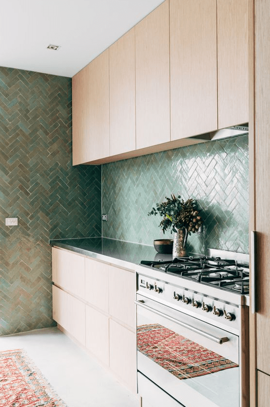 a chic light-stained kitchen with sleek cabinets, a green herringbone tile backsplash and a wall, shiny metal countertops and a cooker