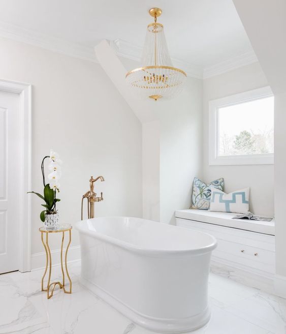 a chic white chandelier with a window, a tub, a chic and glam chandelier, a windowsill bench and a side table with a bloom