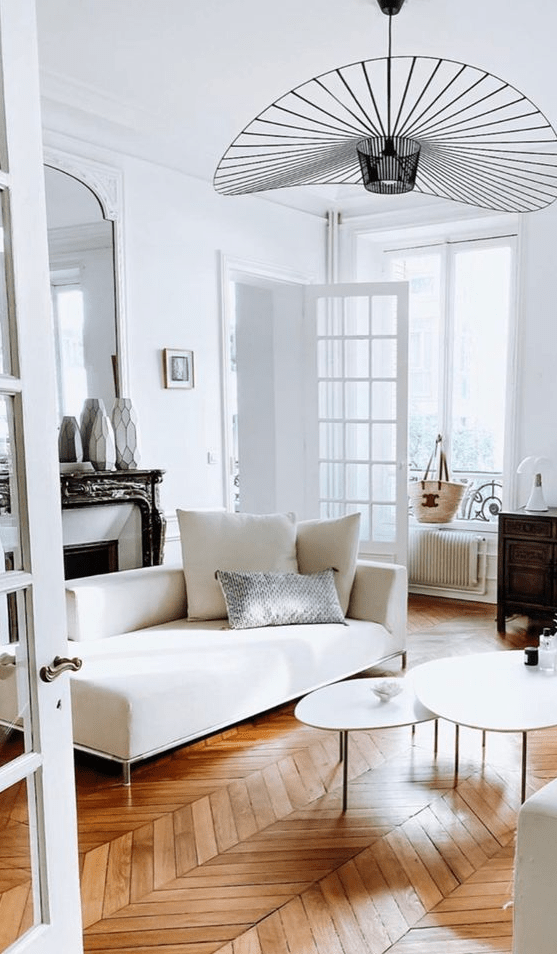 a classy Parisian chic living room with parquet floors, a fireplace, a white daybed, coffee tables and a mirror plus a col lamp