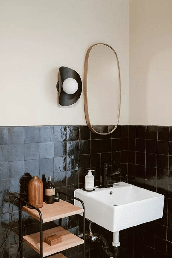 a contrasting bathroom with neutral walls and black Zellige tiles, a tiered shelving unit and a small sink plus a mirror