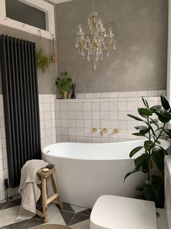 a cool bathroom with concrete walls and white Zellige tiles, a tub, a stool, a crystal chandelier and potted greenery