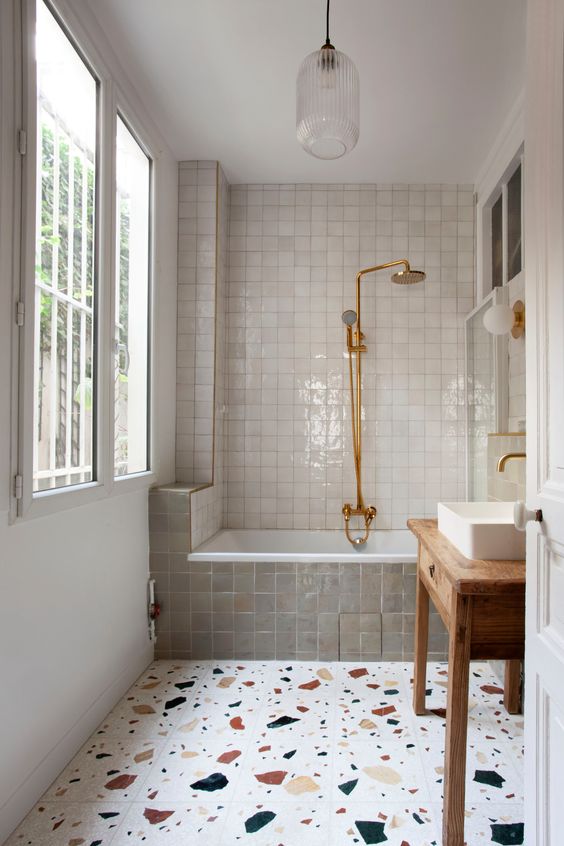 a creative bathroom with a large window, a bath space clad with white and grey Zellige tile, a terrazzo floor, a stained vanity and a sink plus brass hardware