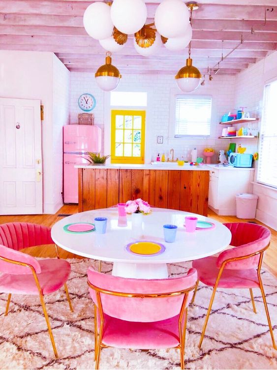 a dopamine decor eat-in kitchen with a pink fridge, colorful tableware, a table with pink chairs and colorful plates