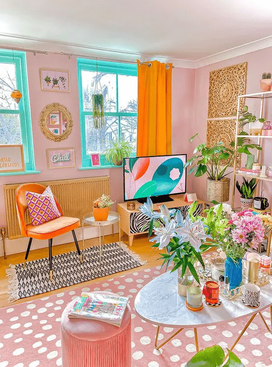 a dopamine decor living room with pink walls, turquoise windows, an orange chair, a pink printed rug, pink pouf and some blooms and greenery